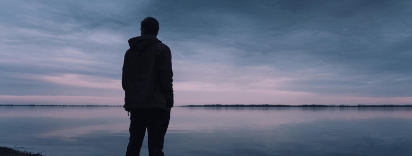 Man in grief stands looking out at water.